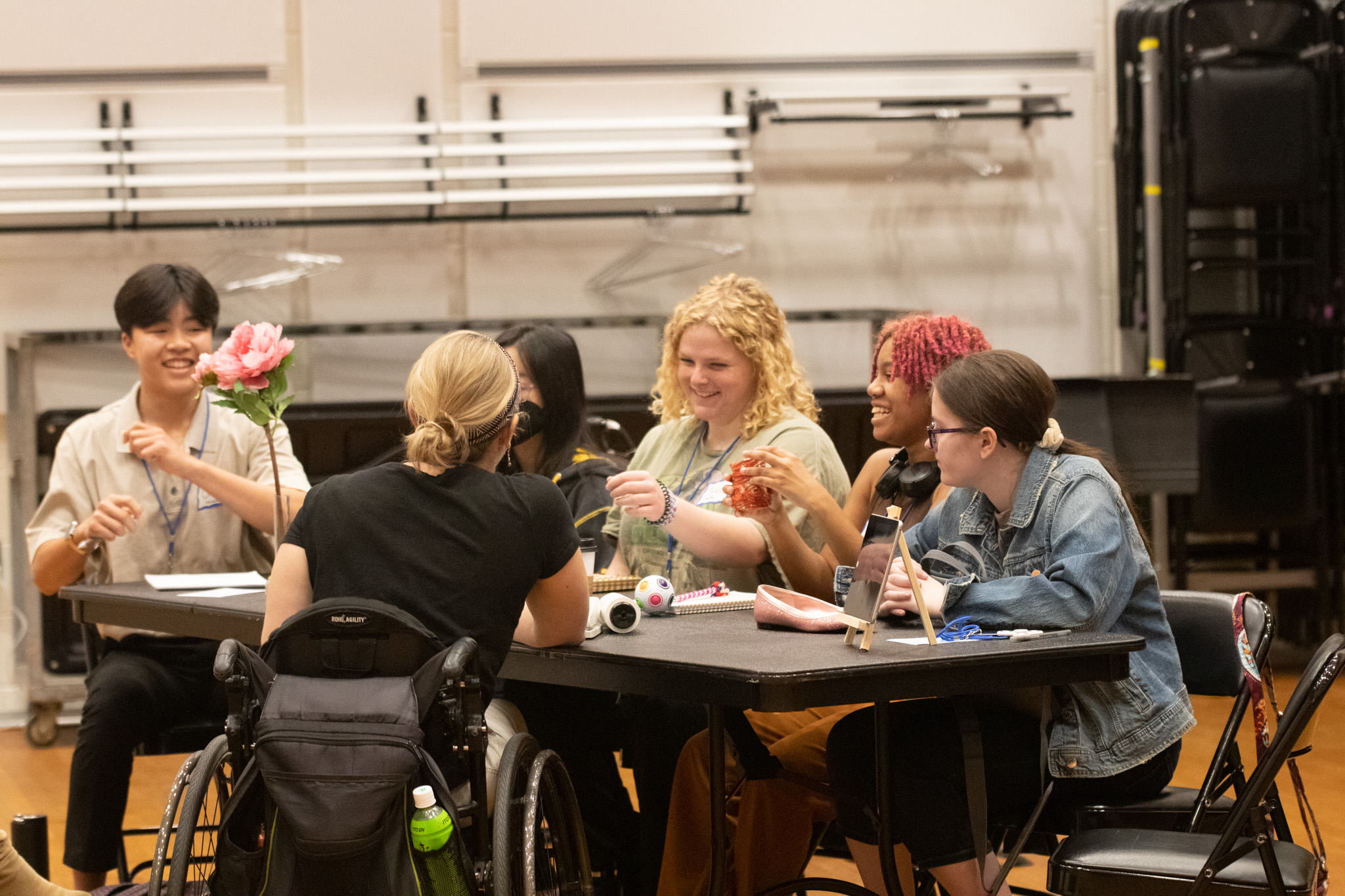 6 people smile as they sit around a rectangular table filled with fidgets and captioning devices.